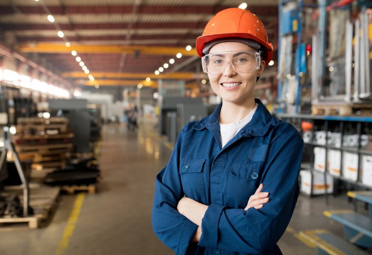 Young smiling female technician in blue uniform, protective eyeglasses and helmet working in modern factory
