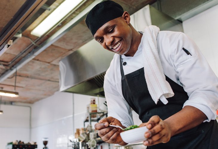 Gourmet chef in uniform cooking in a commercial kitchen. Happy male cook wearing apron standing by kitchen counter preparing food.